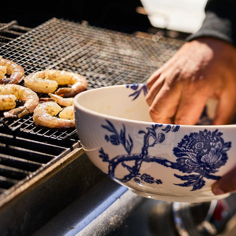 A person is placing seasoned shrimp on a grill using an Arcadia Vegetable Serving Bowl, reminiscent of the elegance found in Caskata Artisanal Home's premium porcelain dinnerware.