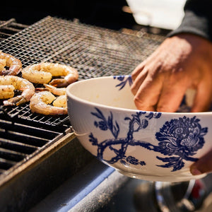 A person is placing seasoned shrimp on a grill using an Arcadia Vegetable Serving Bowl, reminiscent of the elegance found in Caskata Artisanal Home's premium porcelain dinnerware.