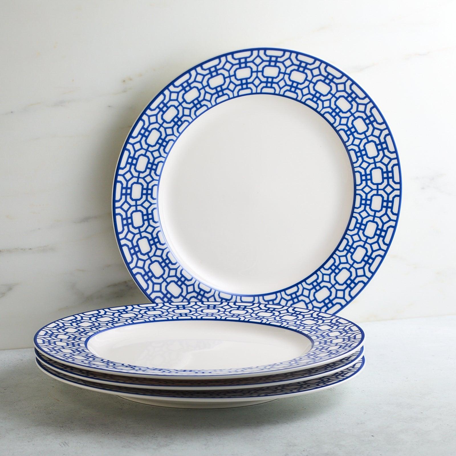 The Newport Garden Gate Table for 4 by Caskata is an elegant porcelain dinnerware set, featuring six dinner plates and six salad plates with blue geometric rims, plus stacked bowls adorned with horizontal blue stripes.