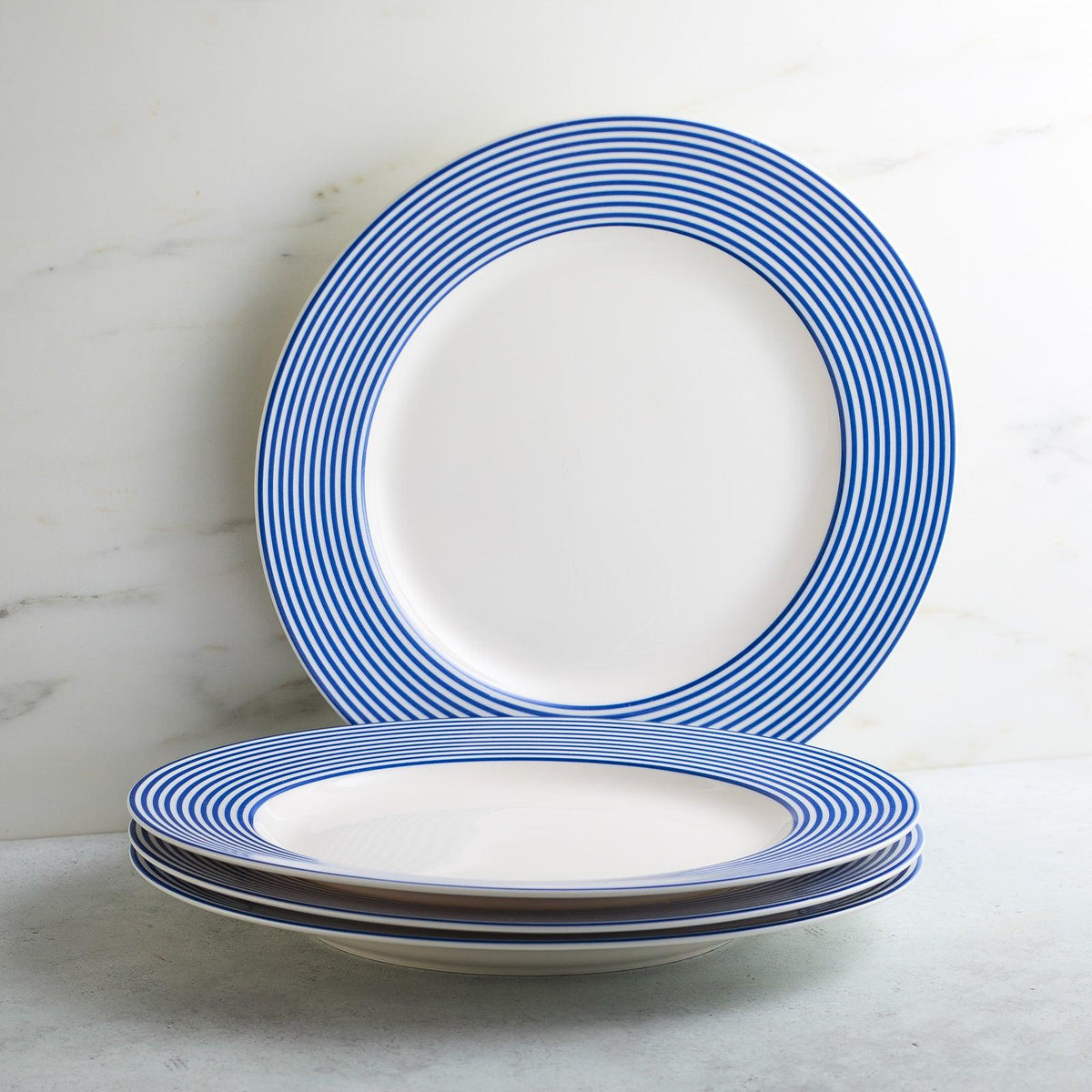 A stack of four Newport Stripe Rimmed Dinner Plates by Caskata Artisanal Home features white bases adorned with blue concentric stripes around the rims. One dinner plate stands upright against the wall behind the stack.