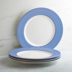 A stack of four Newport Stripe Rimmed Dinner Plates by Caskata Artisanal Home features white bases adorned with blue concentric stripes around the rims. One dinner plate stands upright against the wall behind the stack.