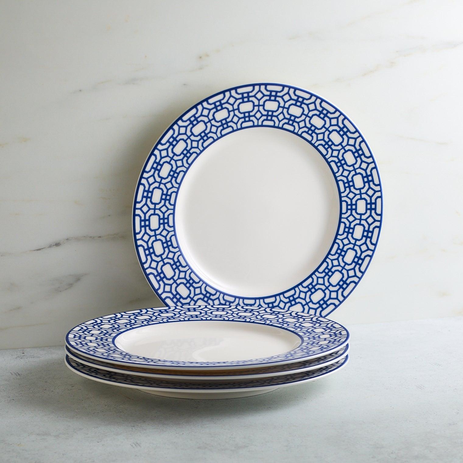 The Newport Garden Gate Table for 4 by Caskata is an elegant porcelain dinnerware set, featuring six dinner plates and six salad plates with blue geometric rims, plus stacked bowls adorned with horizontal blue stripes.