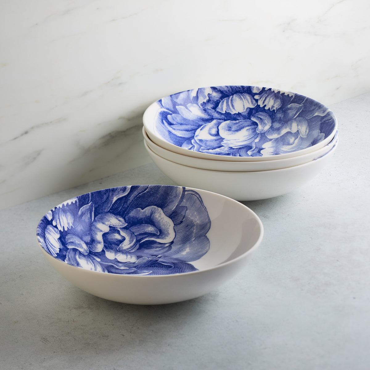Two white Peony Coupe Soup Bowls from Caskata Artisanal Home, with blue peony designs inside, stacked partially on a marble surface.