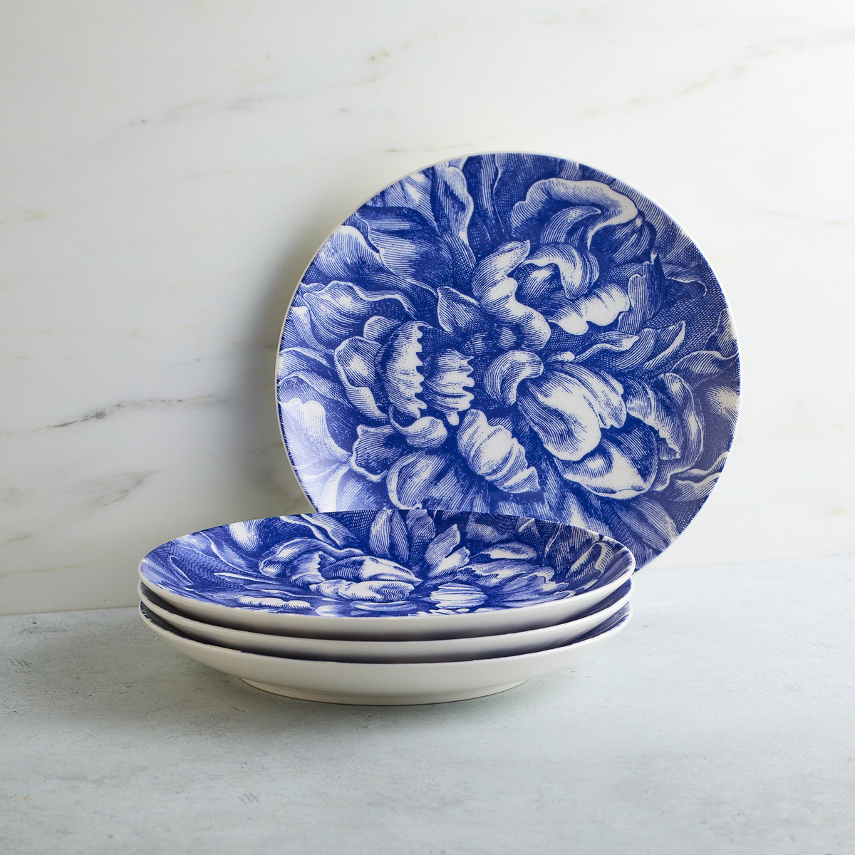 A stack of Caskata Artisanal Home Peony Coupe Salad Plates with a blue floral pattern, positioned against a marble background.