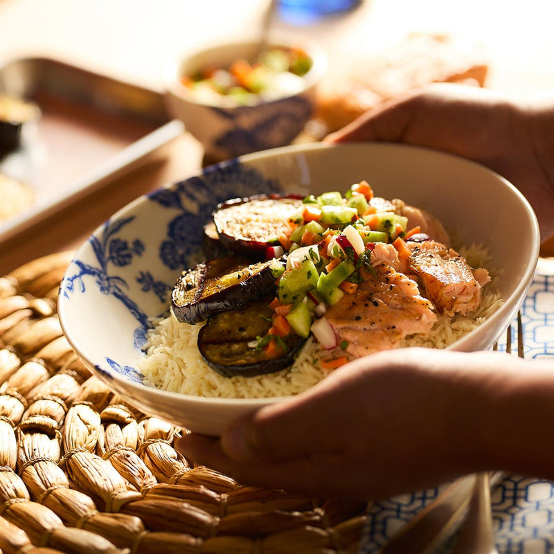 A person holds a Caskata Artisanal Home Arcadia Entrée Bowl of rice topped with grilled salmon, eggplant slices, and diced vegetables.