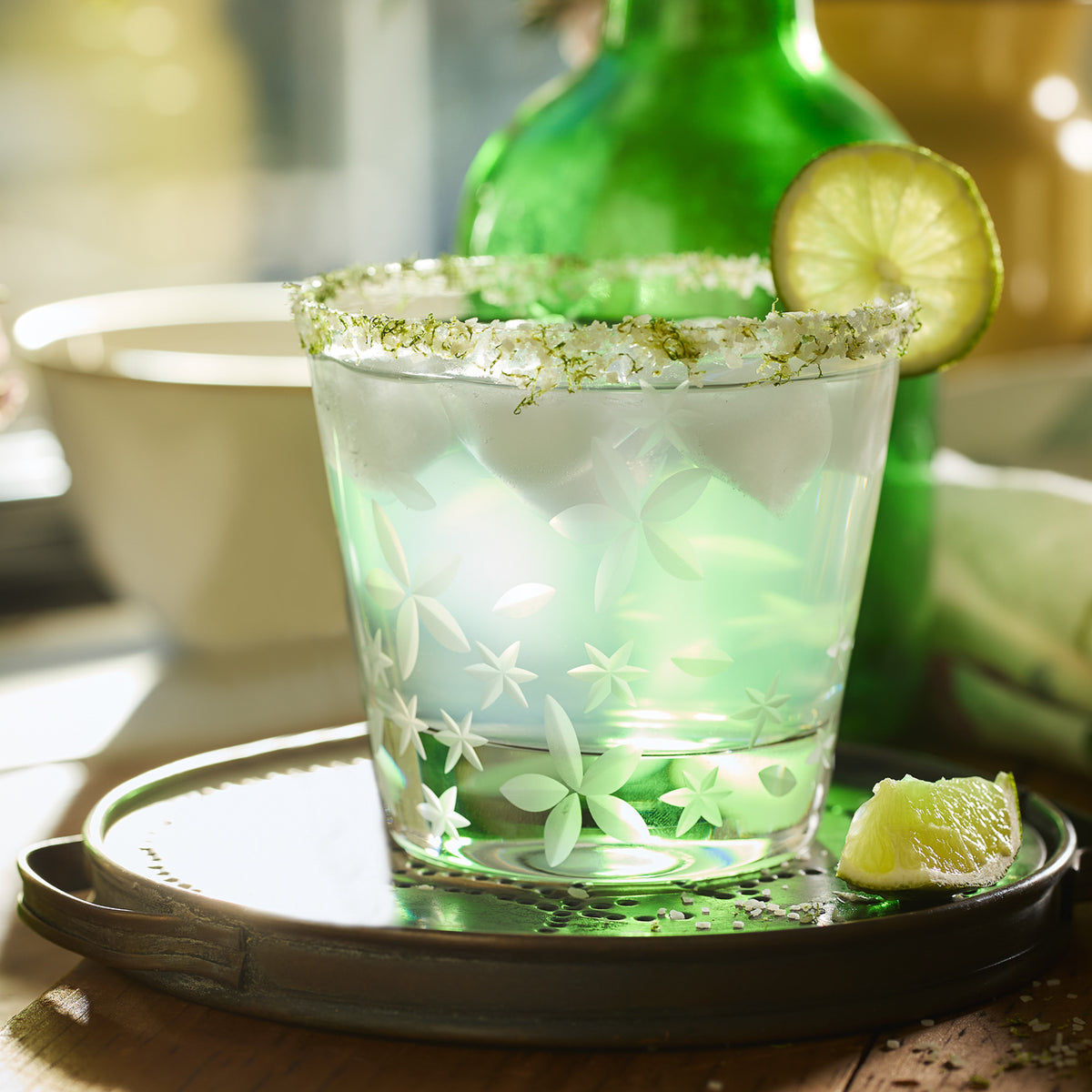 A green-tinted drink with ice in a decorated glass featuring a salt rim and a lime garnish. The lead-free crystal Chatham Bloom Tumblers by Caskata, part of the Chatham Collection, sit on a plate with a lime wedge, with a green bottle in the background. The floral pattern adds an elegant touch to the presentation.