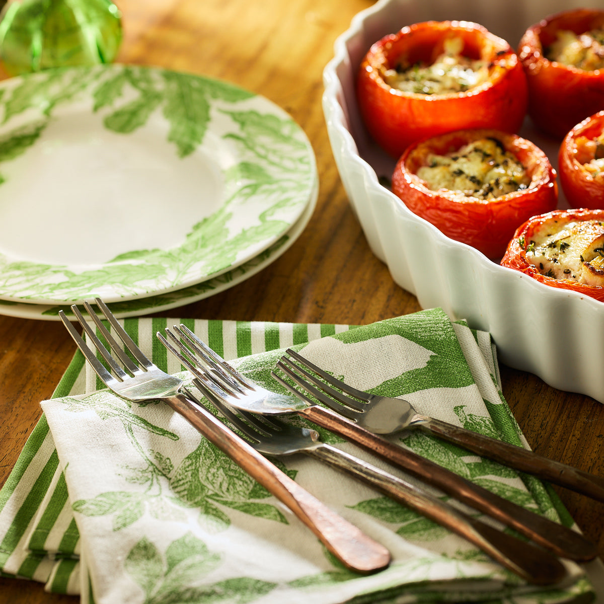 A plate and utensils rest on a Caskata Arbor Birds Verde Dinner Napkin made from 100% cotton, next to a dish of stuffed tomatoes on a wooden table.