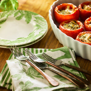 A plate and utensils rest on a Caskata Arbor Birds Verde Dinner Napkin made from 100% cotton, next to a dish of stuffed tomatoes on a wooden table.