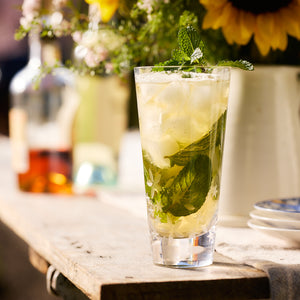 A tall glass of iced tea, filled with ice and fresh mint leaves, sits elegantly on a rustic wooden surface. Crafted from lead-free crystal and part of the Chatham Bloom Highball Glasses by Caskata, the glass adds a touch of sophistication, with blurred bottles and flowers creating a charming backdrop.