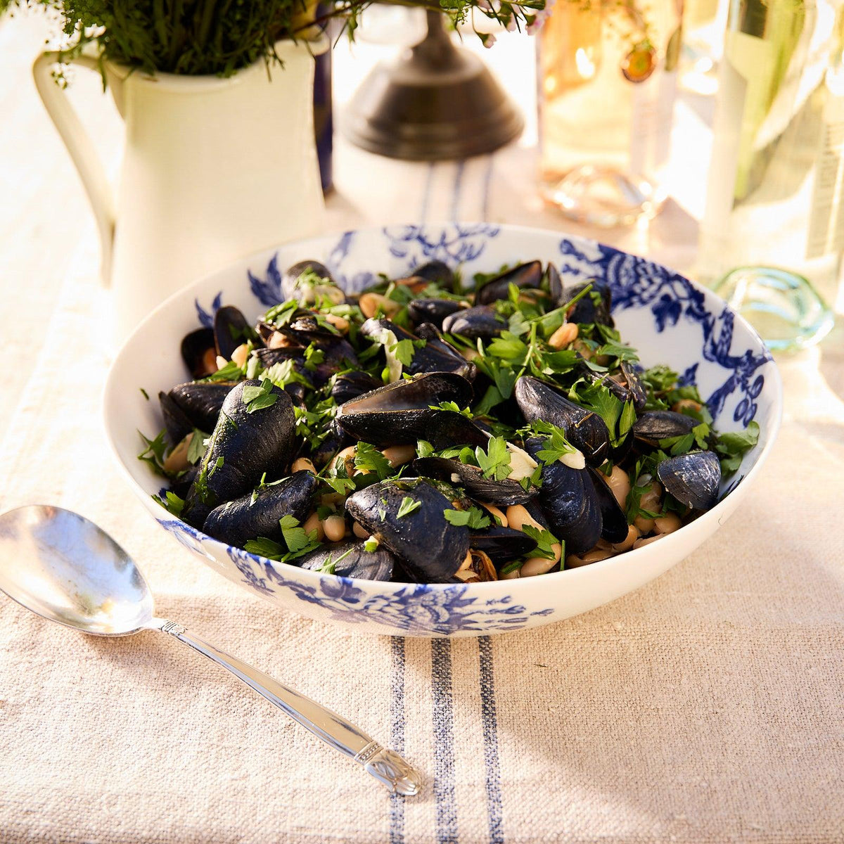 A bowl of cooked mussels garnished with herbs, presented in an Arcadia Wide Serving Bowl by Caskata Artisanal Home, sits on a table adorned with premium porcelain dinnerware. In the background, cutlery and a decorative vase add charm alongside the bottles.