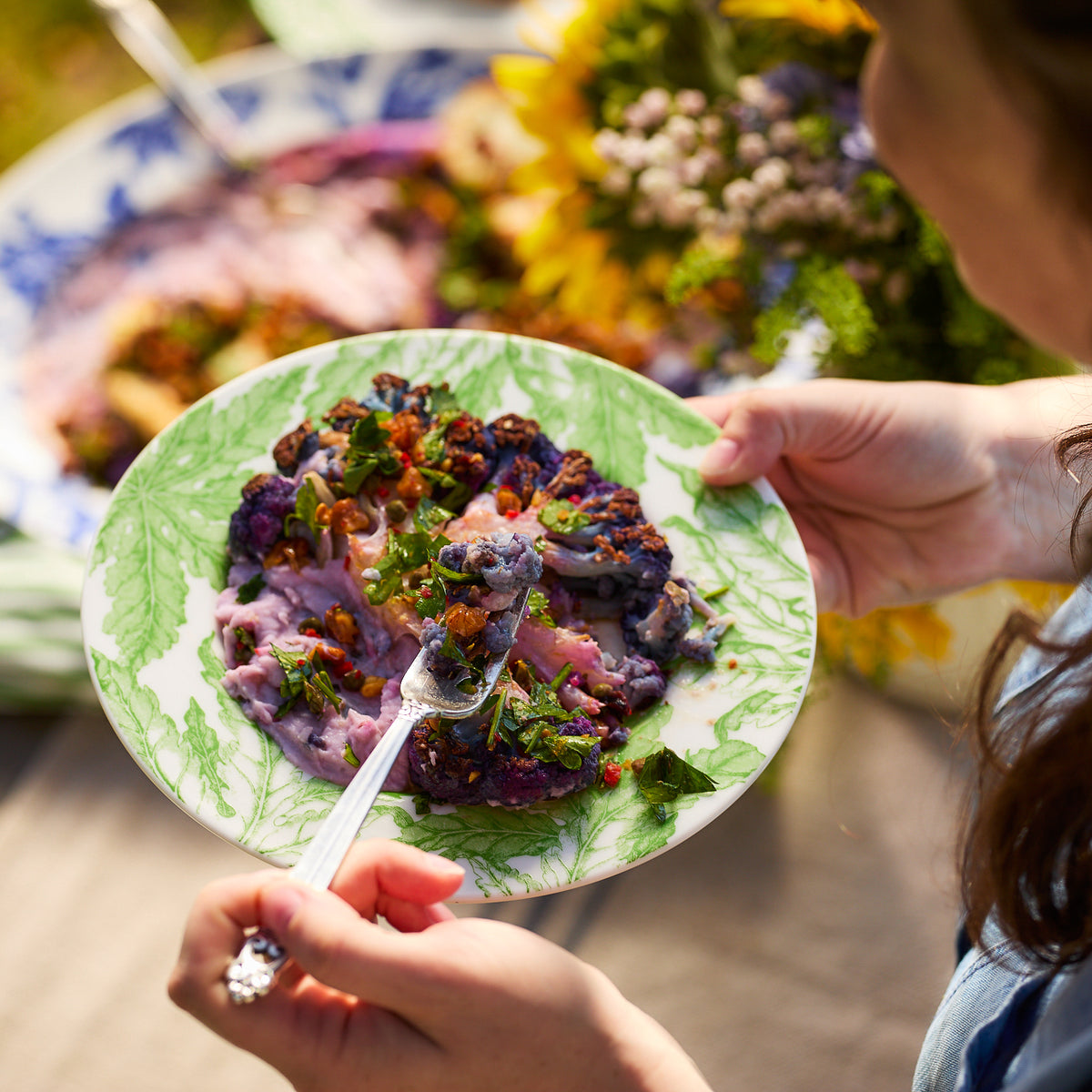 A person holds a Freya Rimmed Salad Plate by Caskata Artisanal Home with purple and green elements, using a fork to eat. A table with more dishes, a bouquet of flowers, and green florals in the background completes the scene.