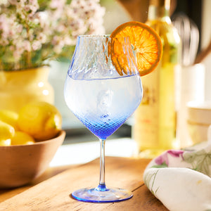 A Caskata Phoebe Cobalt Universal Wine Glass filled with a clear beverage, garnished with a dried orange slice, sits elegantly on a wooden table. In the background, lemons in a bowl and flowers add to the charm.
