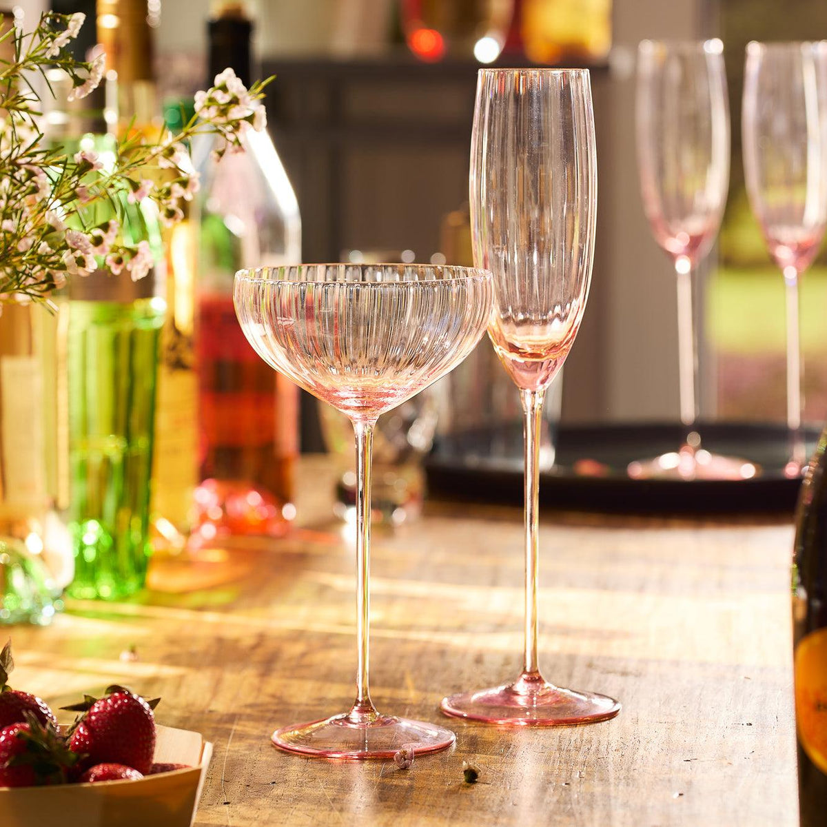 Two elegant Quinn Rose Champagne Flute Glasses by Caskata Artisanal Home, crafted from exquisite lead-free crystal, rest on a wooden table surrounded by bottles and decorations. Mouth-blown in the Czech Republic, they capture the light to create a warm ambiance, with ripe strawberries in the foreground adding a touch of color.
