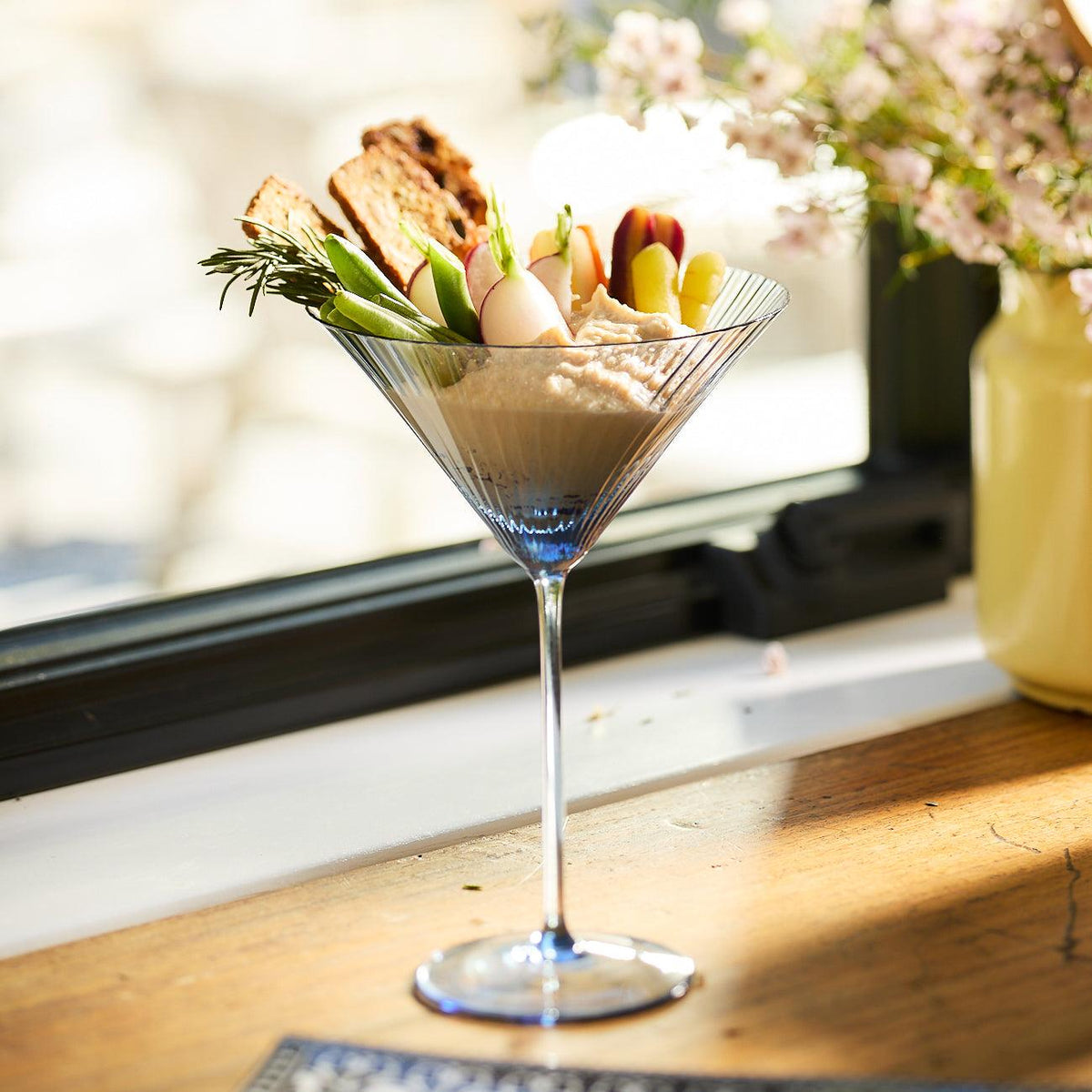 A Quinn Ocean Martini Glass from Caskata Artisanal Home, made of lead-free crystal, filled with a hummus dip, adorned with an assortment of fresh vegetables and crackers, placed on a wooden surface near a window with flowers in a vase beside it.