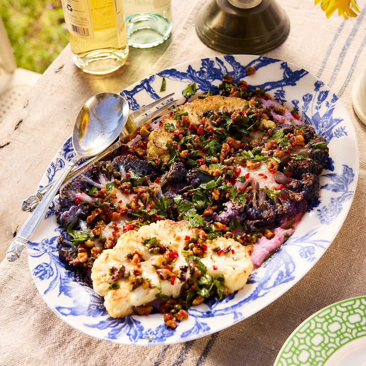 A decorative Arcadia Oval Rimmed Platter from Caskata Artisanal Home contains roasted cauliflower florets garnished with herbs and nuts, accompanied by serving utensils. Bottles and a sunflower are visible in the background, blending elegance and history through Arcadia porcelain dinnerware from the Williamsburg Foundation.
