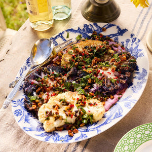 A decorative Arcadia Oval Rimmed Platter from Caskata Artisanal Home contains roasted cauliflower florets garnished with herbs and nuts, accompanied by serving utensils. Bottles and a sunflower are visible in the background, blending elegance and history through Arcadia porcelain dinnerware from the Williamsburg Foundation.