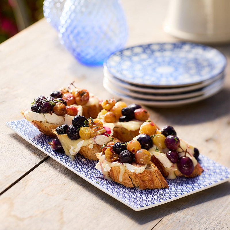 Toasted bread topped with grapes and cheese on a decorative bone china tray create a charming Newport Garden Gate feel. In the background, stacked plates and a textured blue vase complete the scene. Caskata's Newport Petite Serving Tray brings elegance to any setting.
