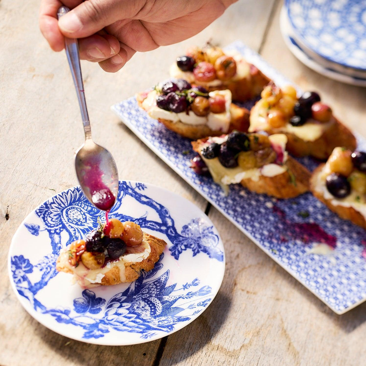 A hand drizzles sauce over a piece of bruschetta topped with grapes and cheese on an heirloom-quality dinnerware plate, with more bruschetta visible on similar Caskata Artisanal Home Arcadia Small Plates in the background.