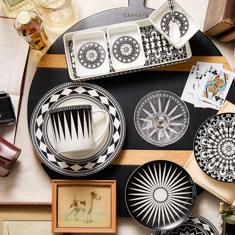 A variety of black and white patterned bone china dinnerware, playing cards, a framed dog illustration, books, and a camera are elegantly arranged on a wooden surface along with the Casablanca Nested Appetizer Tray Set by Caskata.