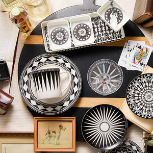 A variety of black and white patterned bone china dinnerware, playing cards, a framed dog illustration, books, and a camera are elegantly arranged on a wooden surface along with the Casablanca Nested Appetizer Tray Set by Caskata.