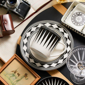 A black and white Marrakech Mug by Caskata Artisanal Home with matching plates featuring Art Deco geometric patterns is placed on a table with a camera, a leather case, framed dog picture, and glassware. The elegant design evokes the timeless allure of Marrakech.