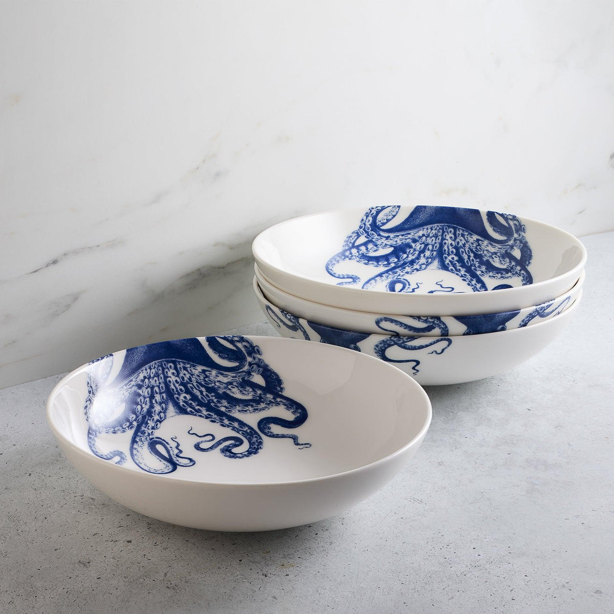 Four premium Lucy Entrée Bowls by Caskata Artisanal Home, each adorned with blue octopus designs, are stacked on a gray surface against a white marble background, evoking a sense of deep-water whimsy.
