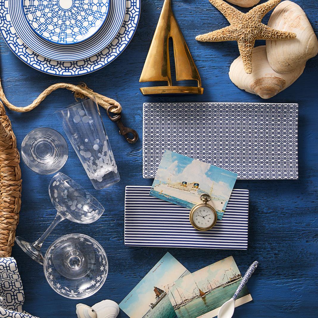 A flat lay featuring Caskata&#39;s Newport Petite Serving Tray with nautical-themed decor, including seashells, a starfish, a brass sailboat, patterned porcelain dishes, postcards, glassware, and a woven basket on a blue wooden surface.