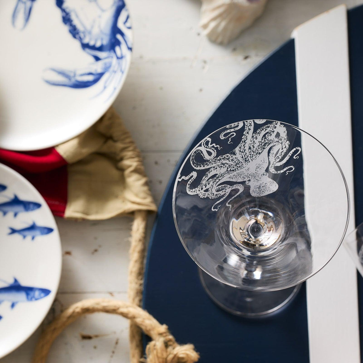 A Caskata Artisanal Home Lucy Martini Glass with an elegant sand-etched octopus design sits next to plates featuring sea creatures on a white and navy table setting.