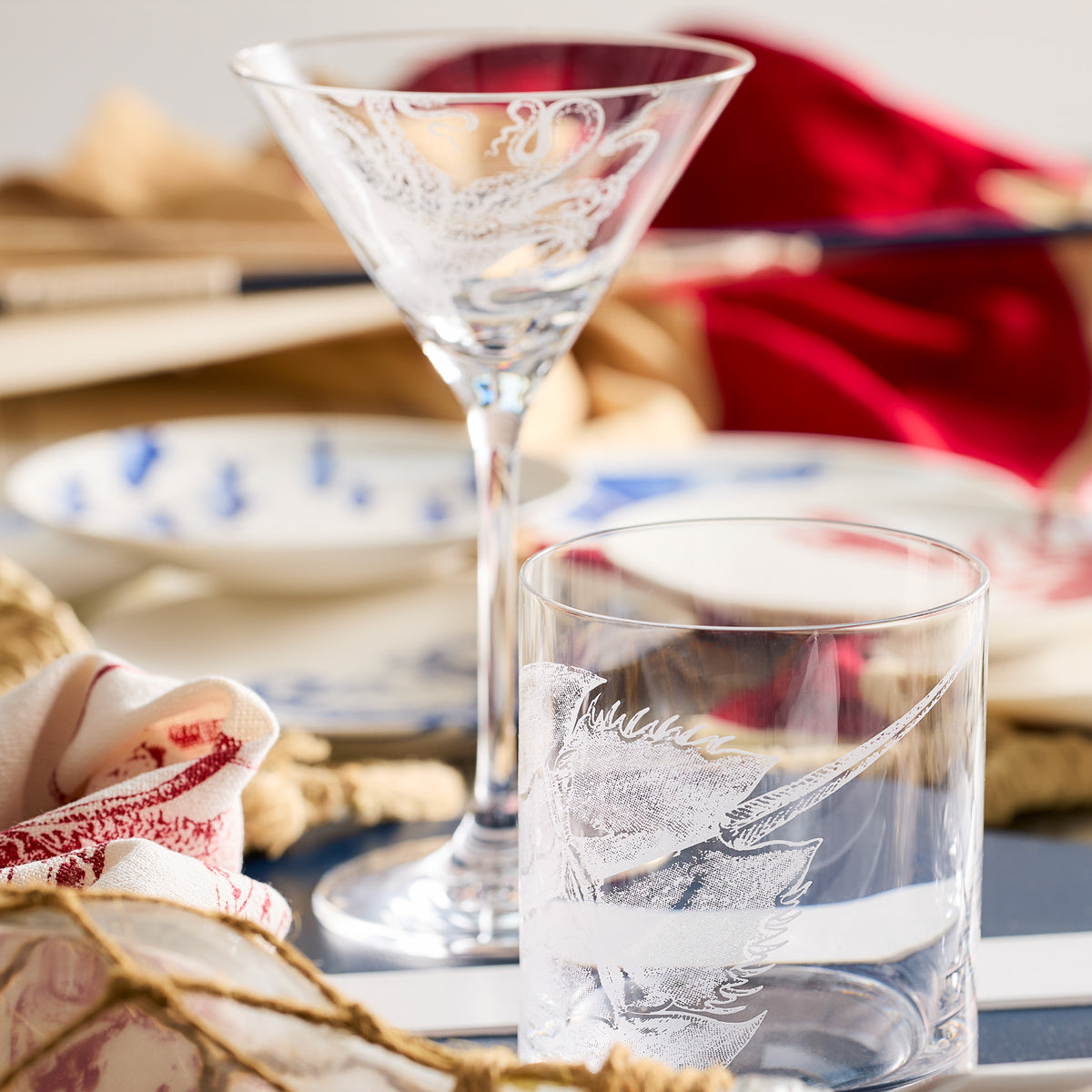 A Caskata Horseshoe Crab Rocks Glass with intricate, sand-etched designs crafted from lead-free crystal graces the table, while plates sit against a rich red cloth in the background.