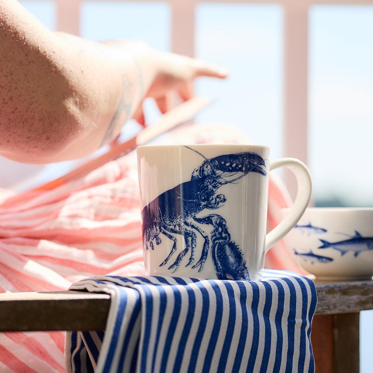 Person pointing while sitting with a blue-and-white striped cloth and a **Lobster Mug by Caskata Artisanal Home** on a wooden table. A bowl with fish design is in the background, adding to the seaside style ambiance.