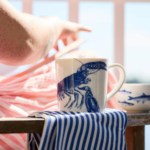 Person pointing while sitting with a blue-and-white striped cloth and a **Lobster Mug by Caskata Artisanal Home** on a wooden table. A bowl with fish design is in the background, adding to the seaside style ambiance.