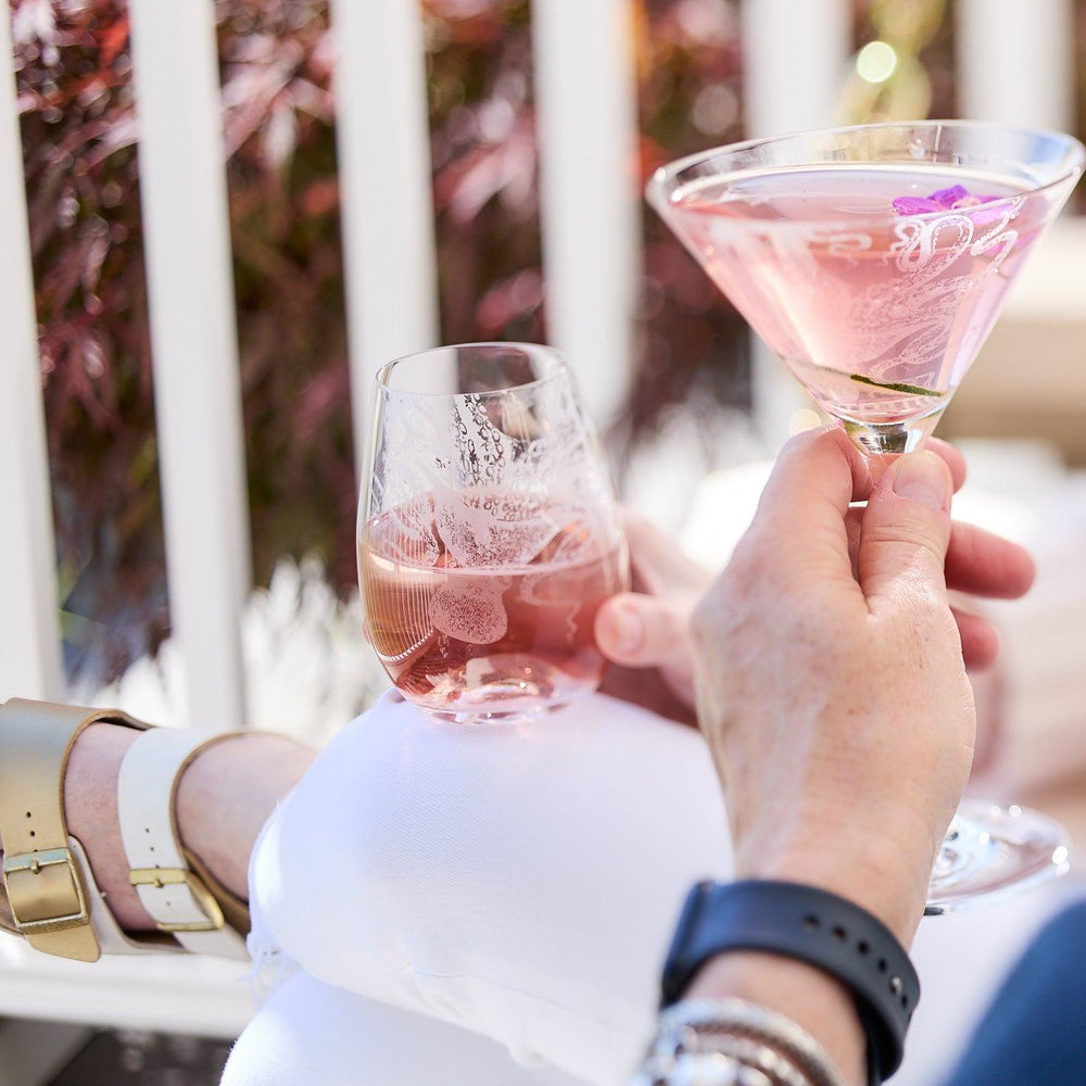 Two people holding drinks, one in a Lucy Stemless Wine Glass by Caskata Artisanal Home and the other in a martini glass, while sitting outdoors near a railing with foliage in the background. One person is wearing a smartwatch and bracelets.