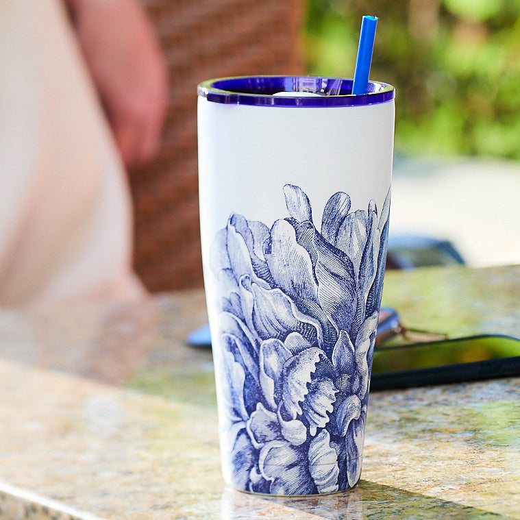 A tall Peony Stainless Steel Insulated Tumbler from Caskata, featuring a white exterior adorned with a blue floral design and accompanied by a blue straw, sits on a granite surface outdoors. This tumbler not only adds to the aesthetic appeal but also boasts vacuum-insulated technology to maintain your beverages at the ideal temperature.