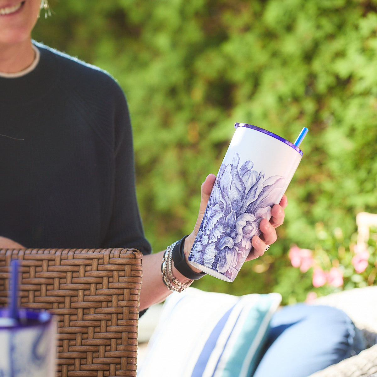 A person is seated outdoors on a wicker chair, savoring their drink from a Caskata Peony Stainless Steel Insulated Tumbler with a blue straw. The stainless steel construction ensures the beverage stays refreshingly cool.