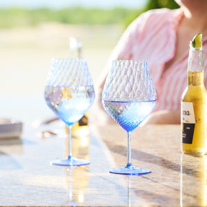 Two playful **Caskata Phoebe Cobalt Universal Wine Glasses** and a clear bottle with a lime wedge rest on an outdoor table. A person in a pink and white striped shirt is partially visible in the background.
