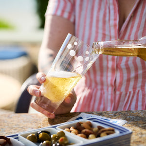 A person in a pink striped shirt pours beer from a bottle into Chatham Pop Highball Glasses by Caskata at an outdoor setting, with a bowl of mixed nuts and olives on the table.