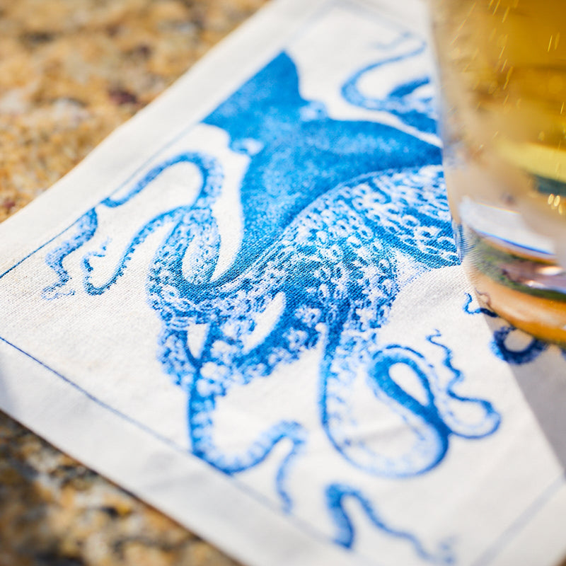 A blue octopus illustration is printed on a Coastal Cocktail Napkin Roll by MY DRAP, showcasing coastal patterns. It's partially covered by a glass with a beverage.
