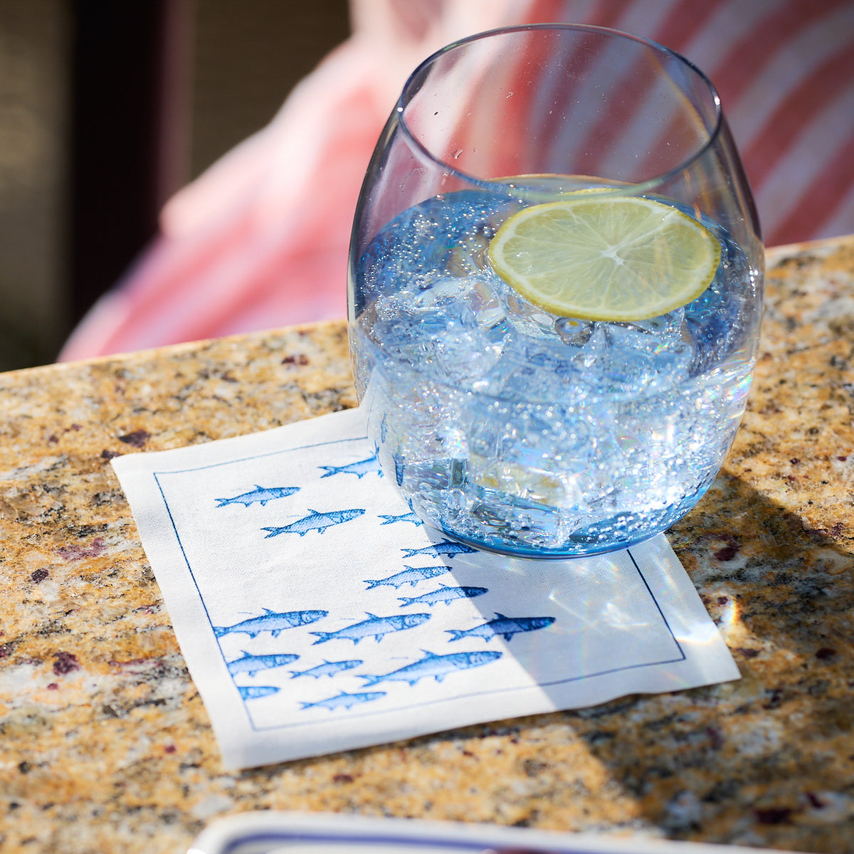 A glass of ice water with a lemon slice rests on an MY DRAP Coastal Cocktail Napkin Roll on a granite countertop.