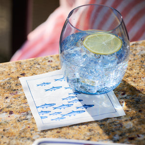 A glass of ice water with a lemon slice rests on an MY DRAP Coastal Cocktail Napkin Roll on a granite countertop.
