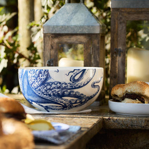 A blue and white ceramic Lucy Large Round Serving Bowl by Caskata Artisanal Home with a Lucy the octopus design is placed on a stone surface, alongside lanterns and a sandwich on a plate. Various food items are partially visible in the foreground.