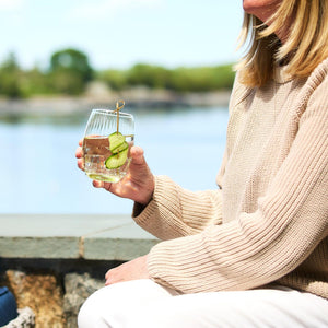 A person wearing a beige sweater holds a glass of water with cucumber slices in a Quinn Citrine Tumbler by Caskata Artisanal Home, sitting by a body of water.