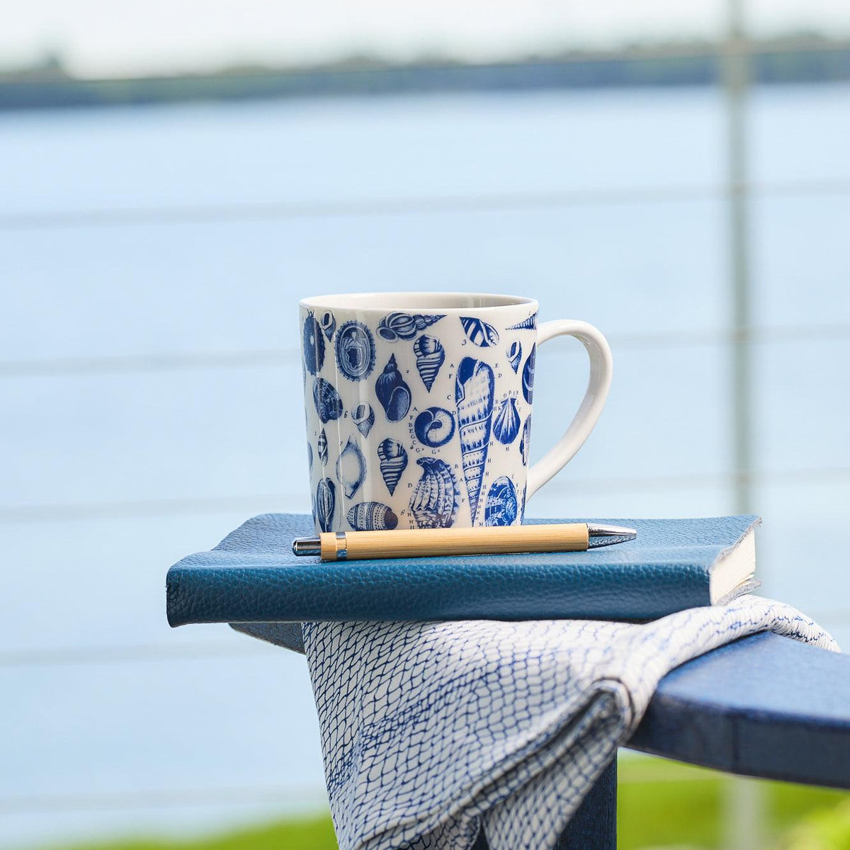 A Shells Mug from Caskata Artisanal Home, featuring a seashell pattern, sits on a blue book with a pen on top. A cloth is draped nearby. The background shows a large body of water and part of a railing. Not only stylish, but it&#39;s also dishwasher and microwave safe.
