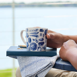 A person holds a generously sized Shells Mug by Caskata Artisanal Home with blue seashell designs while sitting outdoors near the water, with a notebook and pen on their lap.
