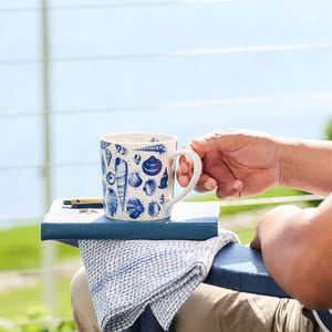 A person holding a Shells Mug from Caskata Artisanal Home, resting on a blue notebook and a checkered cloth. The background shows a blurred view of greenery and water, reminiscent of peaceful beach trips.