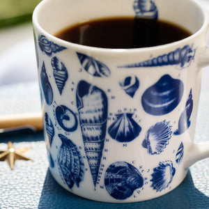 Close-up of a generously sized Shells Mug from Caskata Artisanal Home, made of high-fired porcelain with blue seashell illustrations, filled with black coffee, placed on a blue surface with a small star-shaped object nearby.