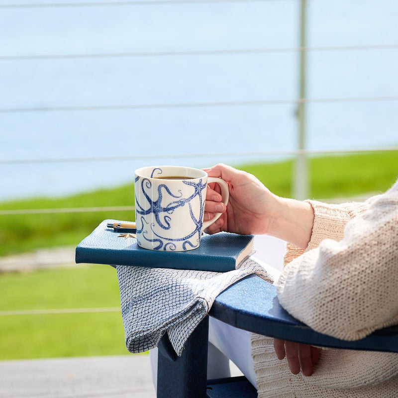 A person holds a high-fired porcelain mug with an octopus design, resting on a chair outside with a body of water and greenery in the background. A notebook and pen sit on the armrest. The Starfish Mug by Caskata Artisanal Home is dishwasher and microwave safe, perfect for enjoying coffee amid nature's tranquility.