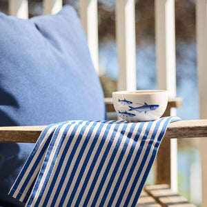 A blue cushion, a striped blue and white cloth, and a small Caskata Artisanal Home School of Fish Snack Bowl are placed on a wooden bench.