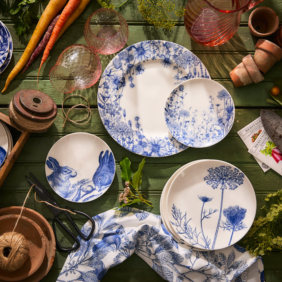A Summer Blues Rimmed Dinner Plate by Caskata Artisanal Home, showcasing a high-fired porcelain with a blue and white floral pattern, adorned on a green table and surrounded by gardening tools, carrots, and glassware.