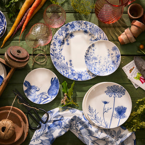A Summer Blues Rimmed Dinner Plate by Caskata Artisanal Home, showcasing a high-fired porcelain with a blue and white floral pattern, adorned on a green table and surrounded by gardening tools, carrots, and glassware.