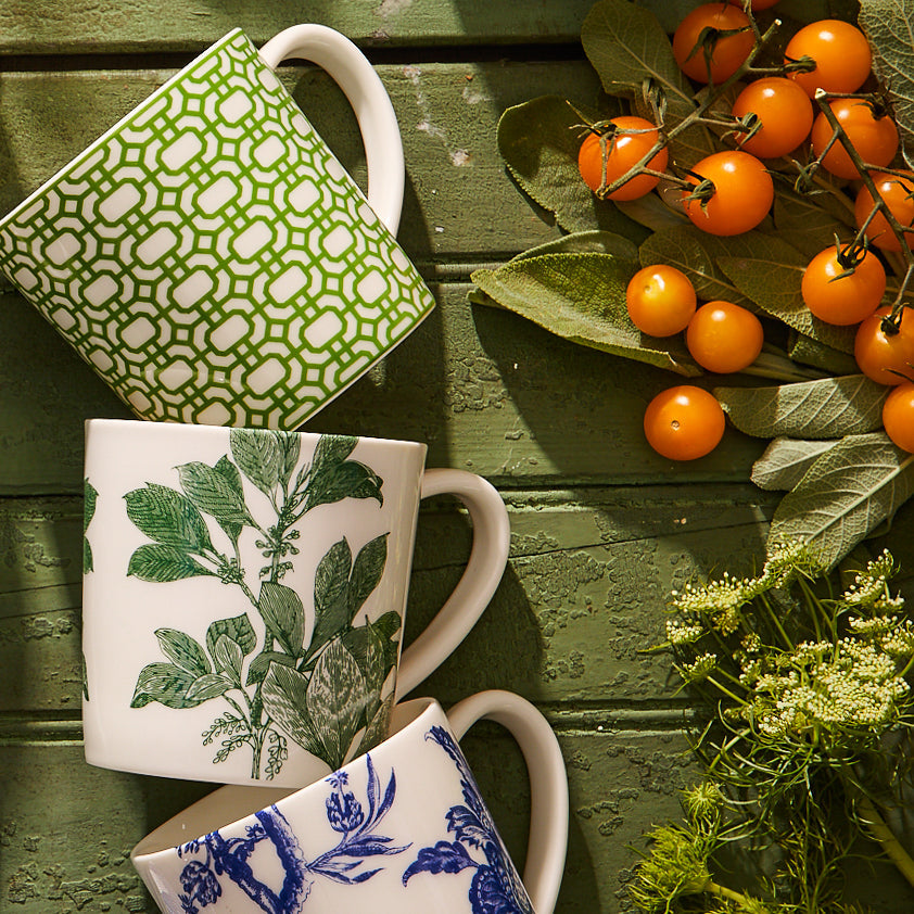 Three Newport Garden Gate Verde Mugs by Caskata Artisanal Home, showcasing green leafy and blue floral patterns, are elegantly arranged on a green wooden surface beside a cluster of yellow berries and green leaves.